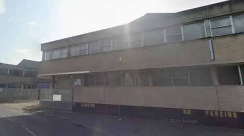 The former Welton Bibby & Baron site. A two-storey building casts a shadow over the road. To the left of the building is an entrance to the site with a metal gate, which is open. 