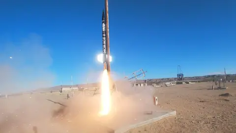 CUSFS A thin black rocket blasting off from a patch of desert. There is a bright light towards the base and blue sky above as it leaves earth. 