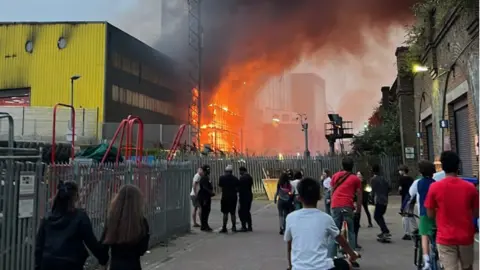 People stand in the foreground looking on as part of a warehouse goes up in flames
