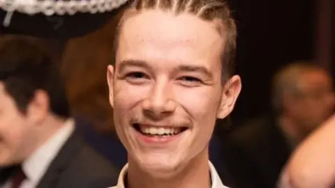 Lizzie Smith A man with his hair arrange in corn rows is smiling and appears to be in a room full of people. He is wearing a white shirt.