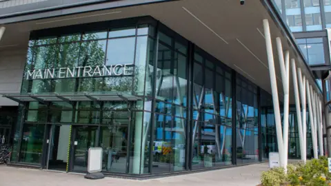 Large glass-fronted entrance to a modern building. There are white support beams and a sign which reads Main Entrance.