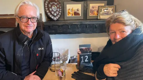 Richard and Carolyn Cater sitting at a table. Richard is wearing glasses and a black jacket. Carolyn has a scarf on. 