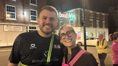 A man with short brown hair and a black sports top beside a woman with blonde hair, glasses and a black top, both standing on a high street with a butchers shop behind them