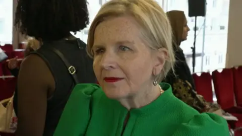 Ockenden in the foreground of what appears to be an indoor event or conference venue. She is wearing a bright green top with structured puffed sleeves and a high neckline. A pearl necklace and small earrings are visible, and she has light-coloured hair styled back from the face.