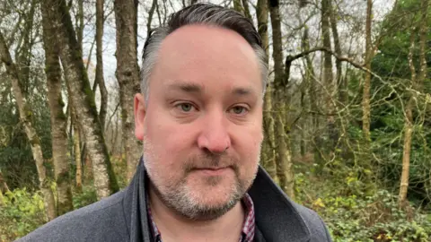 A close-up image of Kenny Donaldson. He is a middle-aged man with slicked back greying hair and a greying beard. He is stood in front of some trees and is looking at the camera with a neutral expression on his face. He is wearing a grey coat with the collar popped and a red checked t-shirt.