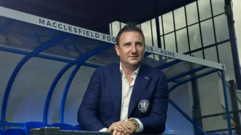 Robert Smethurst wears a navy blue suit with a club logo on the pocket. He wears a white shirt and stands pitch side in front of a a Macclesfield Football Club Home stand.