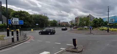 Google Approach to a large roundabout with a sign showing Witton Road in the foreground and a road sign pointing to Witton and Aston Hall. The center of the island has grass, low bushes and a growing tree. Four cars can be seen traveling around the island, and mid-rise buildings can be seen in the background, including one clad in blue tiles.