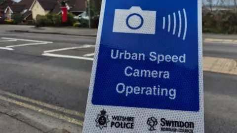 Image of a blue and white temporary road sign which shows an icon of a camera and the words: "Urban Speed Camera Operating" in the background can be seen an urban road, with housing and a red post box.