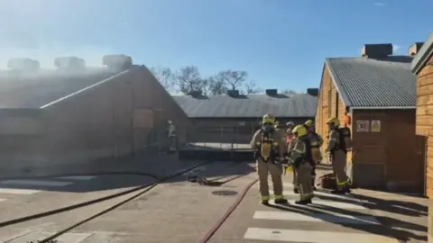 Firefighters stand by buildings on a farm. Some of the buildings look wooden and metal. The firefighters are in unforms. The sun is shining.