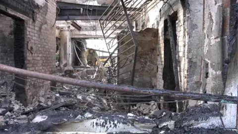 The inside of a home wrecked by fire with rubble on the floor, trees and burnt metal scattered around on the ground.
