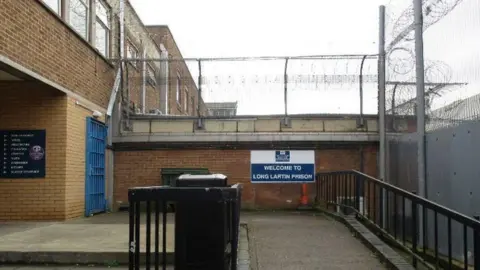 Part of a prison which has a sign saying "welcome to Long Lartin Prison". There are brown walls and a blue gate in the shot. 