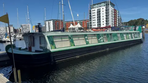 George King/BBC A large green barge floating on water at a marina in front of blocks of apartments.