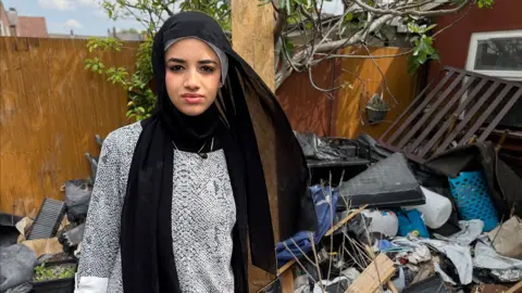 Razan, in a black head scarf, stands in her back garden surrounded by compost, seed trays and cannabis growing detritus