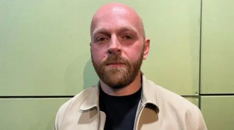 Sean Egan is looking at the camera, in front of a green wall. He is bald and has a beard, and is wearing a light-beige jacket with a black T-shirt.