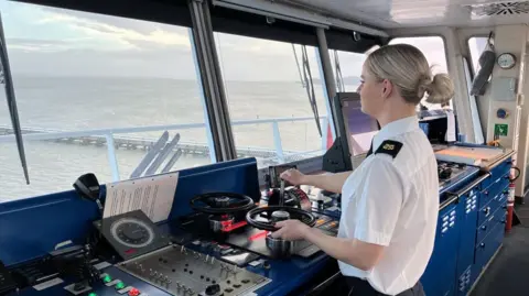 Lauren Lucas helming Wight Sky car ferry. She is in uniform with a white shirt and black epaulettes. She has her back to the camera and is looking out towards the Solent.