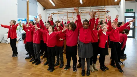 Lucy Walters Children rehearsing their performance in what appears to be a dancing studio. They have their hands in the air. They are wearing red uniforms.