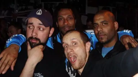 Kaptin Barrett Four people pose closely together for a photograph in a dimly lit indoor venue. One person stands in the foreground at the centre of the group, while three others gather behind and beside them. The image is tightly cropped around the group, with blue sports-style tops visible on two people in the background and dark clothing on others.