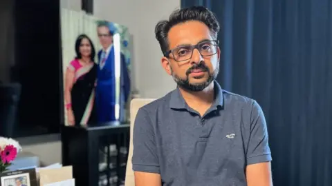 Miten Patel poses for a picture. He wears glasses and wears a blue polo shirt. A portrait of what appears to be his parents is seen in the background of the room.