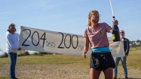 RWREDMANPHOTOGRAPHY A young woman wearing a pink top and black shorts is visibly happy and laughing while holding a large bottle of wine having just finished a run. There are two people behind her holding aloft a white banner that says 204 days 200 marathons on it