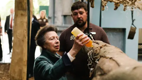 Robbie Dark Photography Princess Anne is squeezing an amber-coloured liquid onto a branch. A man is watching her.