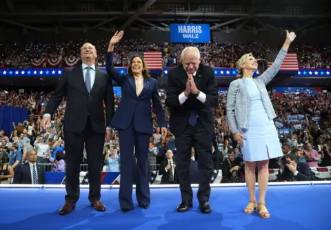 Getty Images Doug Emhoff, Kamala Harris, Tim Walz and Gwen Walz wave at a crowd at a rally