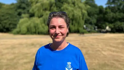 Amanda Hill, wearing a blue T-shirt, with a logo on it, standing in a field, with trees behind it. She is smiling and has short dark hair, with sunglasses on her head.