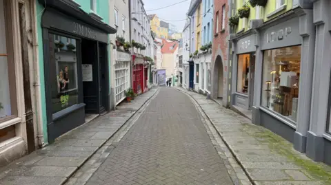 A shot of Mills Street. There are shops either side of a cobblestone road and two people walking side by side in the distance.  