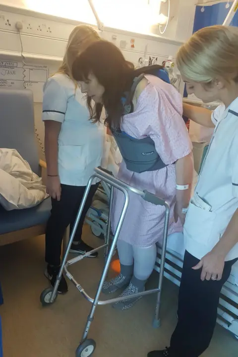 Dave Mathias A woman is using a walking frame. Two medical staff stand next to her
