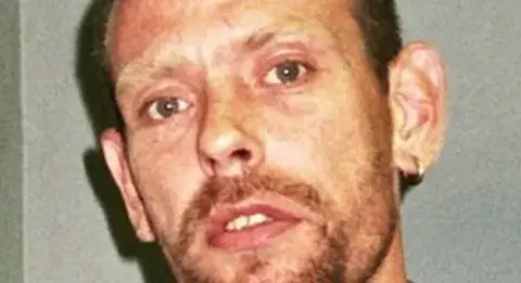 Suffolk Police A close-up image of Paul Clarke. He is looking directly into the camera and standing in front of a grey wall.