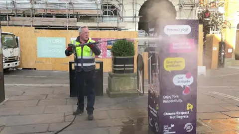 A man in a hi-vis vest jet-washes street furniture in York city centre.