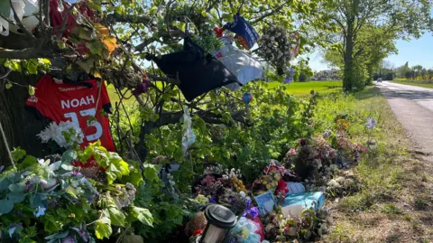 Tony Fisher/BBC Flowers and tributes laid by a road in memory of Noah. There is an orange Luton Town football club shirt hanging off a branch with his name on it and the number 13.