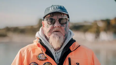 BBC Angler Jay Lewis who has a white beard, facial tattoos and a septum nose ring is wearing a grey cap, orange waterproof jacket and is looking at the camera 