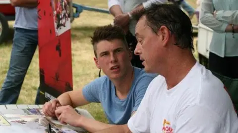Sam Coulter A man in a white t-shirt sits next to his teenage son, wearing a blue t-shirt in a field, surrounded by people. They are at a table while others look on.