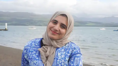 Jacob Lloyd/MFL Mentoring Head and torso shot of Firial Benamer, who is a woman of Libyan heritage in her early twenties. She is wearing a light tan headscarf tied in front of her neck and a light blue jacket with a pattern of different white leaves in a loose arrangement of squares across it. She is wearing light pink lipstick and has her head tilted to the side and is smiling directly at the camera. She is standing on a beach of either the sea or a lake, with hills in the distance and three small boats visible but slightly out of focus behind her.
