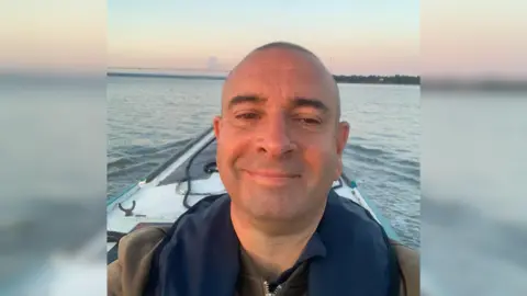 A man stood on a boat taking a selfie of himself. The sea is behind him and it is sunset.