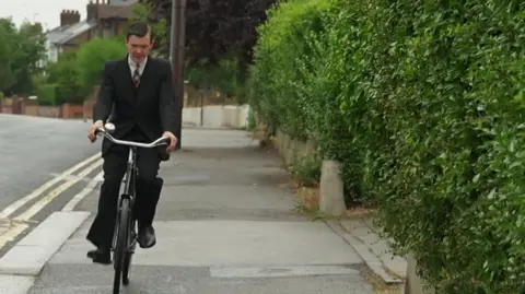 Jodie Halford/BBC Lincoln rides on old style push bike along a pavement that is lined with bushes.