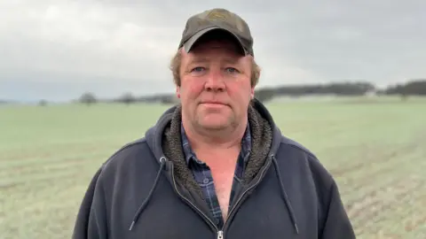 Dan Willis stands in a field at his farm. He is wearing a khaki coloured cap and a check blue and navy shirt under a zipped hooded jumper. The field behind him is tinted white with frost. 