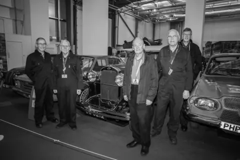 Günter and Christine Starke Five male Coventry Transport Museum volunteers stand amid vehicles at the site. Most are wearing dark overalls with lanyards around their necks. 