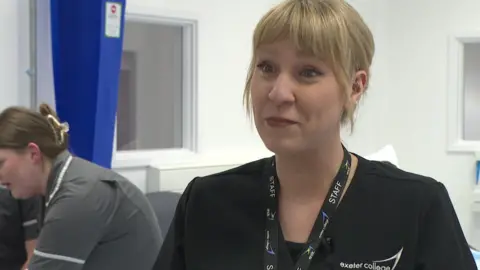 The image shows a member of healthcare teaching staff at Exeter College speaking in a simulated hospital ward, wearing a staff lanyard and uniform with the college logo visible. In the background, students in clinical uniforms are practising on a training bed, reinforcing the hands-on learning environment.
