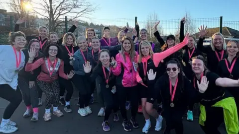Runclub Members of the club are gathered together for a group photograph. They are all wearing running gear and have pink lanyards and medals. There is a fence in the background with winter trees and a blue sky.