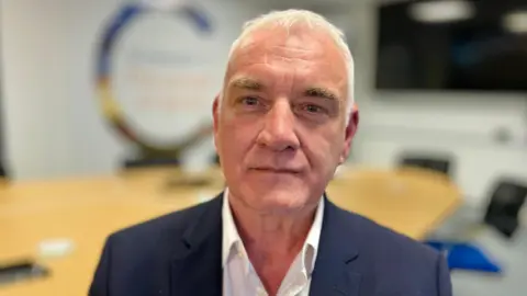 Martin Giles/BBC A man with grey hair looks at the camera. He is wearing a white open-necked shirt and a navy suit jacket 