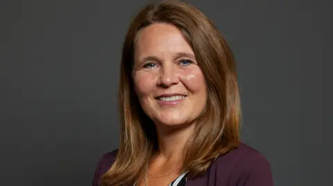 Official Parliamentary portrait of MP Vikki Slade. She has shoulder length chestnut-brown hair and is wearing a deep-purple jacket.