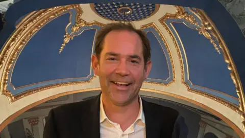 James Mews A smiling man is standing inside the Opera House.
He is near the a blue and gold dome in the ceiling.
