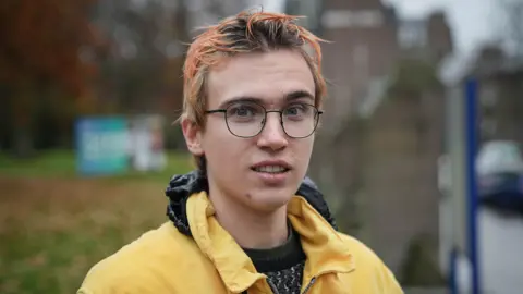 James Elliot has short, light brown and orange-tinted hair and is wearing glasses and a yellow jacket. He is standing outdoors on a cloudy day with blurred buildings and autumn trees in the background.