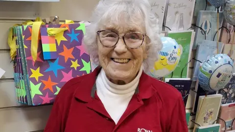 A bright and smiley elderly lady wearing a red polo shirt stands next to a display of cards and badges