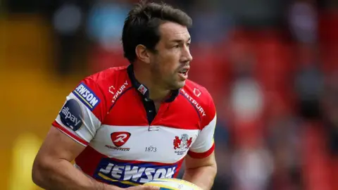 Action Images/Reuters Tom Voyce running with a rugby ball during a professional match. He has dark hair and wears a mouth guard, and is looking away from the camera. He also wears a red and white Gloucester Rugby shirt. He is in focus, but the rest of the background is heavily blurred. 