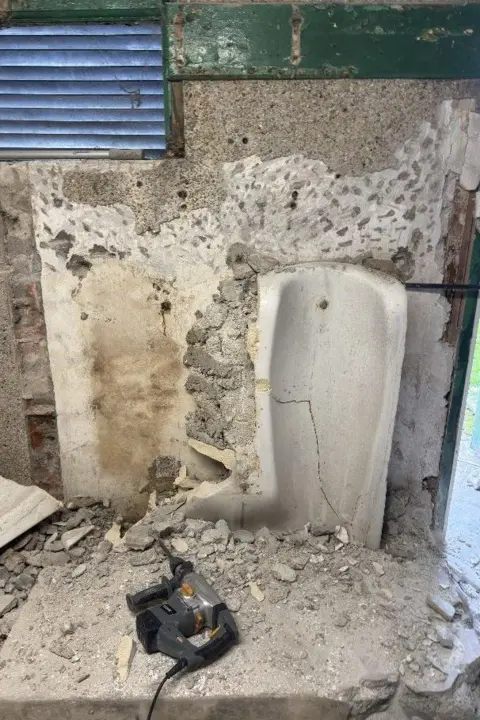 Ellie Pizey A cracked urinal in the derelict building with an electric drill laid next to it. There is broken plaster everywhere and there is a small window which has a shutter across it.