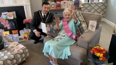 Edith Hill, surrounded by birthday cards, is reading one from the King and Queen. She is wearing a light blue dress and pink sash and tiara