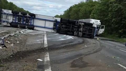A white and blue lorry lies on it's side across the central reservation with all the wheels and tyres showing and dirt, dust and white paint marks across the road.