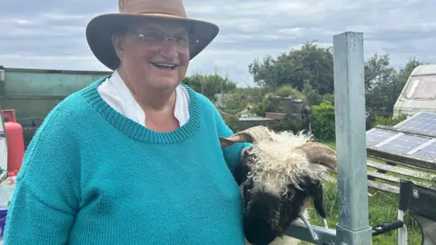 A woman wearing a turquoise jumper and a brown hat stroking a sheep